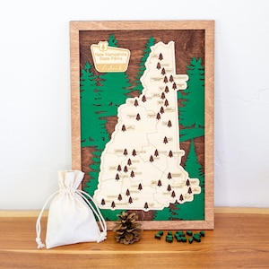 May include: Wooden map of New Hampshire State Parks with a green forest background and a wooden frame. The map features small tree icons marking park locations. A small drawstring bag, pine cone, and miniature trees are in front of the map.
