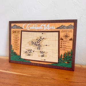 May include: Wooden map of Colorado with the text "Colorado 14ers" in a cursive font. The map highlights 58 peaks over 14,000 feet. The frame is brown with green and brown accents, and the text "The mountains are calling and I must go."
