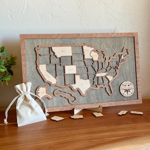 May include: A wooden wall map of the United States featuring light-colored state cutouts on a gray background within a brown frame. A circular compass emblem is attached to the right, and a small white drawstring pouch sits nearby.