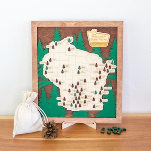 May include: Wooden framed map of Wisconsin State Parks with a light wood frame, green trees, and a light-colored map. The map has small wooden tree markers. A small bag, pine cone, and wooden stand are in front of the map.