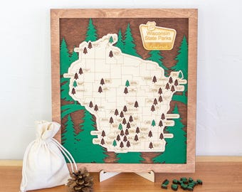 Pode incluir: Um mapa emoldurado em madeira dos parques estaduais de Wisconsin com um recorte de mapa de madeira clara sobre um fundo verde escuro com silhuetas de pinheiros. O mapa inclui pequenos ícones de árvores e uma placa com Wisconsin State Parks.
