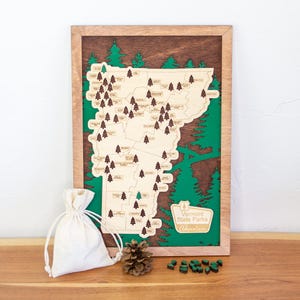 May include: Wooden map of Vermont State Parks with a brown frame, featuring a light wood map with green accents and small wooden tree markers. A small white bag, pine cone, and wooden trees are on the wooden surface.