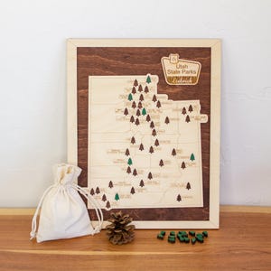 May include: A rectangular wooden map of Utah State Parks featuring a dark brown background with a light wood map overlay. The map includes small tree icons, a title plaque reading Utah State Parks, a small beige drawstring bag, and a pinecone.