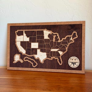 May include: A rectangular wooden wall map of the United States featuring a dark brown background and a lighter wood frame. The map includes cut-out state shapes, with some states in light wood and others in dark wood. A circular compass rose emblem reads "Adventure Awaits The United States of America."