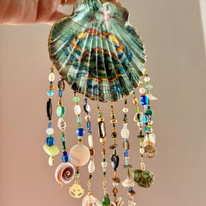 May include: A decorative seashell wind chime with a painted design in shades of blue, green, and gold. The chime features strands of colorful beads, shells, and charms, including a pineapple and a tree of life charm.