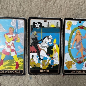 May include: Three tarot cards are displayed, featuring detailed illustrations. The "Page of Swords" card shows a figure with a sword. The "Death" card depicts a cloaked figure on horseback. The "the WORLD" card shows a nude figure within a wreath.