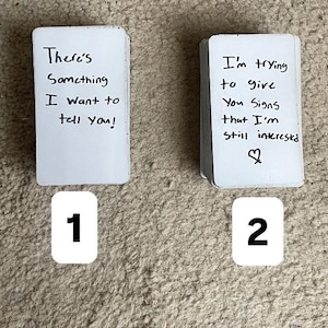 Könnte beinhalten: Zwei Stapel weißer rechteckiger Karten mit abgerundeten Ecken. Der erste Stapel zeigt den handschriftlichen Text: There's Something I want to tell you! Der zweite Stapel zeigt den handschriftlichen Text: I'm trying to give You Signs that I'm still interested mit einem Herzsymbol.