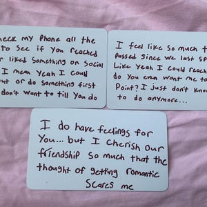 May include: Three white cards with handwritten text in black ink. The cards have different messages about feelings and relationships. The background is a pink fabric.