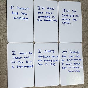以下が含まれることがあります： 手書きの青いインクで書かれたテキストが書かれた6枚の白いカード。カードには「すべてを話したわけではない」や「あなたにロマンチックな興味はない」などのフレーズが含まれています。カードはニュートラルな色調の表面に配置されています。