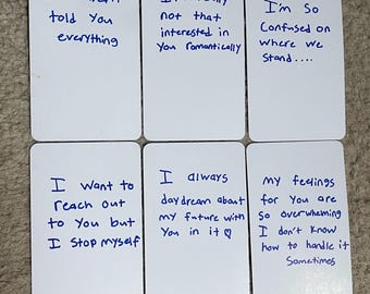 Puede incluir: Seis tarjetas blancas con texto manuscrito en tinta azul. Las tarjetas contienen frases como "No te lo he contado todo" y "Realmente no estoy interesado en ti románticamente". Las tarjetas están dispuestas sobre una superficie de tono neutro.