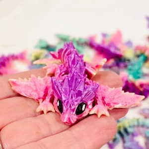 May include: A small, pink and purple dragon figurine with detailed scales and large eyes. The dragon is made of a smooth, glossy material and has a whimsical, fairytale-like appearance.