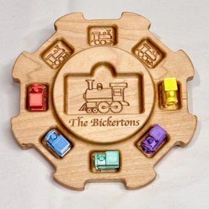 May include: Wooden train-themed toy with a central train engraving and the text "The Bickertons." The toy features a circular design with colourful miniature train cars in red, yellow, blue, purple, and teal.