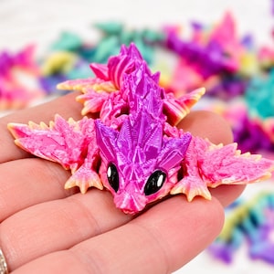 May include: A small, articulated dragon figurine featuring a vibrant gradient of bright pink and deep purple. The creature has a textured, crystalline appearance with sharp, pointed scales along its back, wide wings, and large, glossy black eyes.