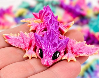 May include: A small, articulated dragon figurine featuring a vibrant gradient of bright pink and deep purple. The creature has a textured, crystalline appearance with sharp, pointed scales along its back, wide wings, and large, glossy black eyes.