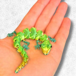 Peut inclure: Un jouet articul&eacute; en forme de gecko vert et or. Le gecko est pos&eacute; sur le ventre, les pattes tendues. Le jouet est en plastique et a une apparence r&eacute;aliste.