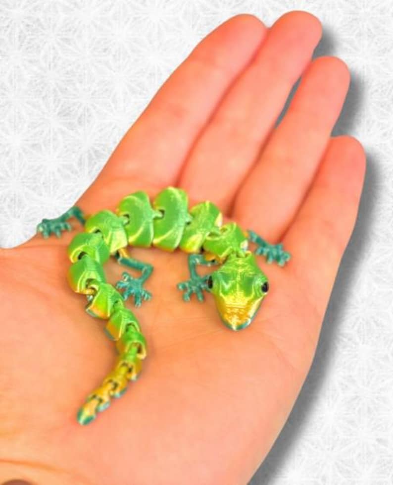 Peut inclure: Un jouet articul&eacute; en forme de gecko vert et or. Le gecko est pos&eacute; sur le ventre, les pattes tendues. Le jouet est en plastique et a une apparence r&eacute;aliste.
