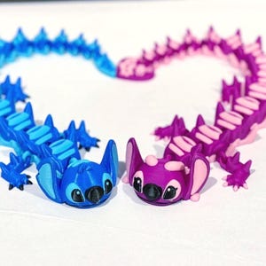 Peut inclure: Deux figurines articulées, une bleue et une violette, disposées en forme de cœur. La figurine bleue a un accent bleu clair, et la figurine violette a un accent rose. Les deux figurines ont de grands yeux noirs et sont posées sur une surface blanche.