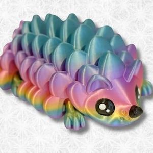 May include: A rainbow-colored 3D printed hedgehog figurine with a cute face and large eyes. The figurine is made of plastic and has a smooth, glossy finish.