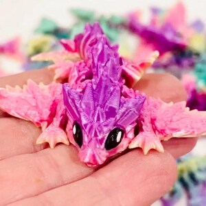 Peut inclure: Une petite figurine de dragon rose et violette avec des ailes. Le dragon a une surface textur&eacute;e d&eacute;taill&eacute;e et de grands yeux noirs.