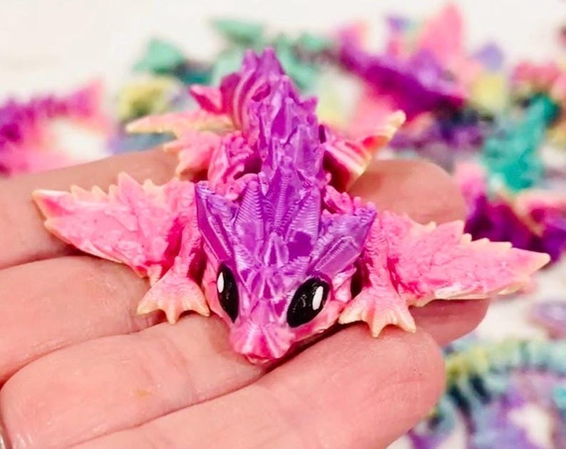 Peut inclure: Une petite figurine de dragon rose et violette avec des ailes. Le dragon a une surface textur&eacute;e d&eacute;taill&eacute;e et de grands yeux noirs.