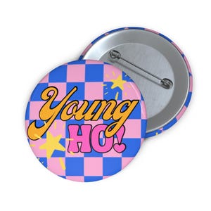 Pode incluir: Um broche redondo com um padrão xadrez azul e rosa. O texto Young HO! está impresso numa fonte estilizada laranja e rosa. Gráficos de estrelas amarelas estão espalhados pelo design e um mecanismo de alfinete de metal é visível.