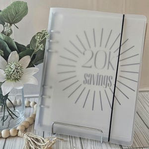 May include: A clear binder with the text "20K savings" in gray, surrounded by a sunburst design. The binder is held in place by a black elastic band. The binder is displayed on a metal stand.