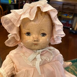 May include: A vintage doll with a heavily cracked, pale face, blue eyes, and painted red lips. The doll wears a light pink fabric bonnet with ruffled edges and a matching pink dress featuring white lace trim and a tied ribbon at the neck.