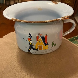 May include: A light blue enamel potty with a hand-painted design of a boy in a red hat and blue uniform walking towards a yellow house with red chimneys. The house has a rooster on the roof and two trees in front of it.