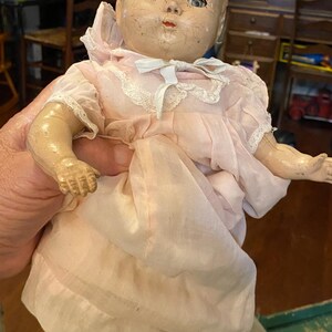 May include: A vintage composition doll with a cracked face, blue eyes, and painted features. The doll wears a pale pink dress with short puffed sleeves, white lace trim around the collar, and a small white ribbon bow at the center of the neckline.