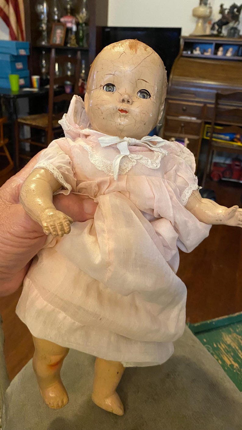 May include: A vintage composition doll with a cracked face, blue eyes, and painted features. The doll wears a pale pink dress with short puffed sleeves, white lace trim around the collar, and a small white ribbon bow at the center of the neckline.