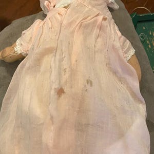 May include: A vintage doll wearing a pale pink fabric bonnet and a matching long, sheer, light pink dress with visible signs of wear, including small stains and frayed edges. The doll has light-colored fabric arms and a soft, draped appearance.