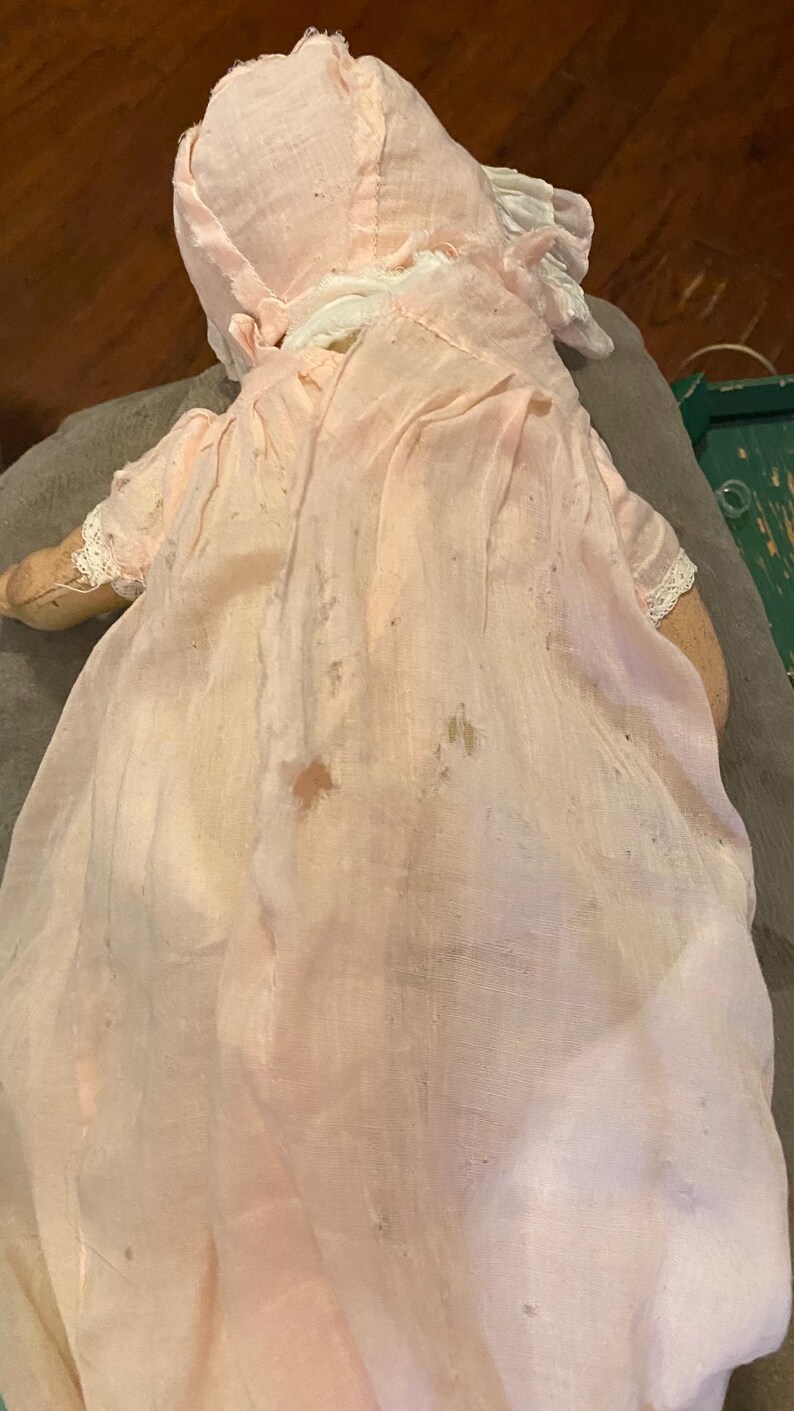 May include: A vintage doll wearing a pale pink fabric bonnet and a matching long, sheer, light pink dress with visible signs of wear, including small stains and frayed edges. The doll has light-colored fabric arms and a soft, draped appearance.