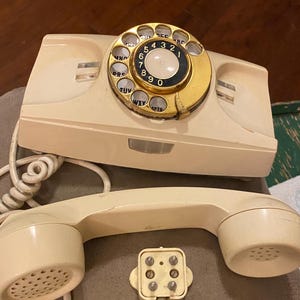 May include: A vintage rotary dial phone with a cream-colored body and a gold-colored dial. The phone has a separate handset with a cream-colored body and a black speaker. The phone has a six-prong plug.