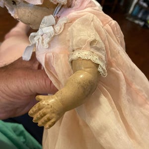 May include: A vintage doll with a cracked face and tan skin, wearing a pale pink bonnet and a matching pink dress with white lace trim on the sleeves and collar. The doll has visible signs of wear, including dark spots on the arm and fabric.