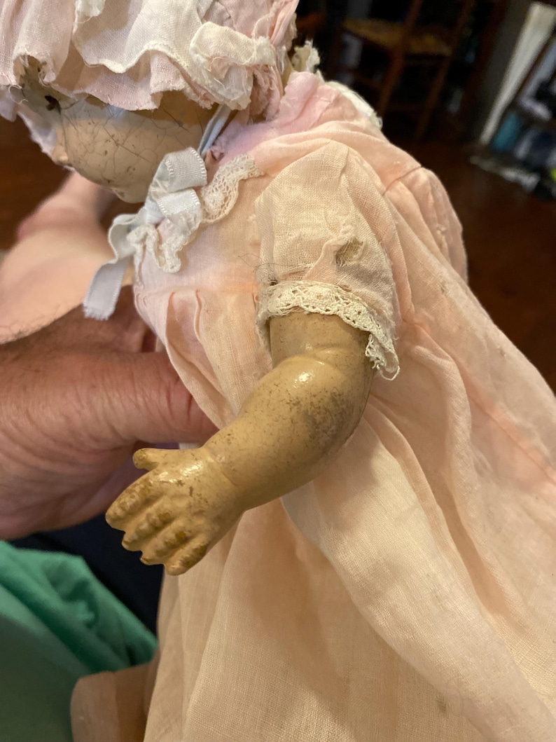 May include: A vintage doll with a cracked face and tan skin, wearing a pale pink bonnet and a matching pink dress with white lace trim on the sleeves and collar. The doll has visible signs of wear, including dark spots on the arm and fabric.