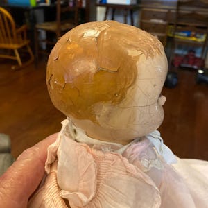 May include: A vintage doll with a cracked, light-colored head and painted brown hair. The doll features a visible network of fine cracks across the surface of the head and face, and it is dressed in a light pink, ruffled fabric garment with white lace trim.