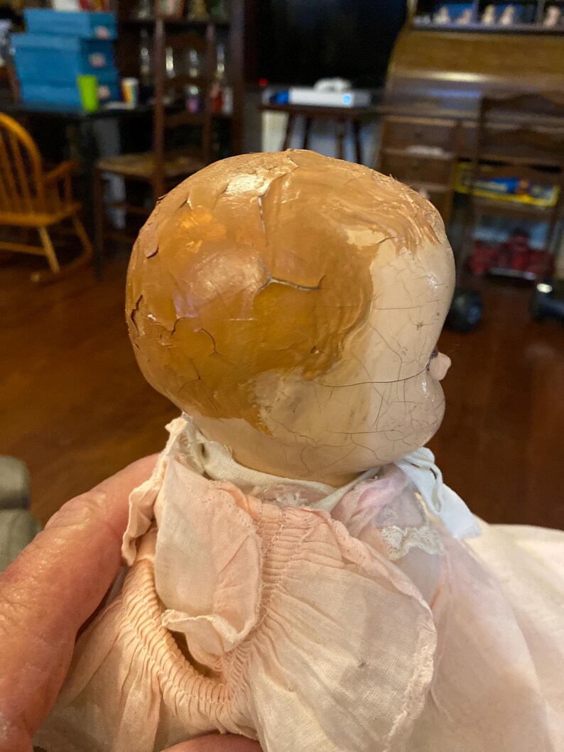 May include: A vintage doll with a cracked, light-colored head and painted brown hair. The doll features a visible network of fine cracks across the surface of the head and face, and it is dressed in a light pink, ruffled fabric garment with white lace trim.