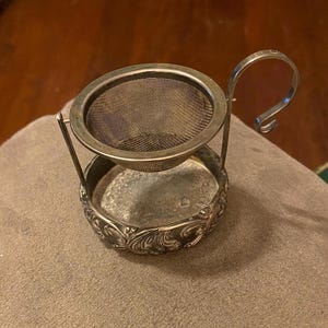 May include: Silver tea infuser with a mesh strainer and a decorative base. The infuser has a handle that can be used to lift it out of a teapot.