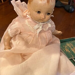 May include: A vintage composition doll with a cracked, light-toned face, painted blue eyes, and molded hair. The doll wears a pale pink, long-sleeved dress featuring a ruffled collar, white lace trim, and a white ribbon tie at the neck.