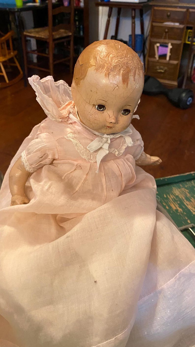 May include: A vintage composition doll with a cracked, light-toned face, painted blue eyes, and molded hair. The doll wears a pale pink, long-sleeved dress featuring a ruffled collar, white lace trim, and a white ribbon tie at the neck.