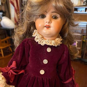 May include: A doll with light brown hair and painted facial features, wearing a long-sleeved, deep burgundy velvet dress. The dress features a beige lace collar, three white buttons down the front, and beige lace cuffs with small burgundy bows.