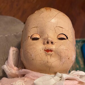 May include: A doll with a pale, cracked composition head featuring closed eyes, painted red lips, and a small nose. The doll wears a light pink fabric garment with a white lace trim collar and a tied white ribbon bow at the neck area.