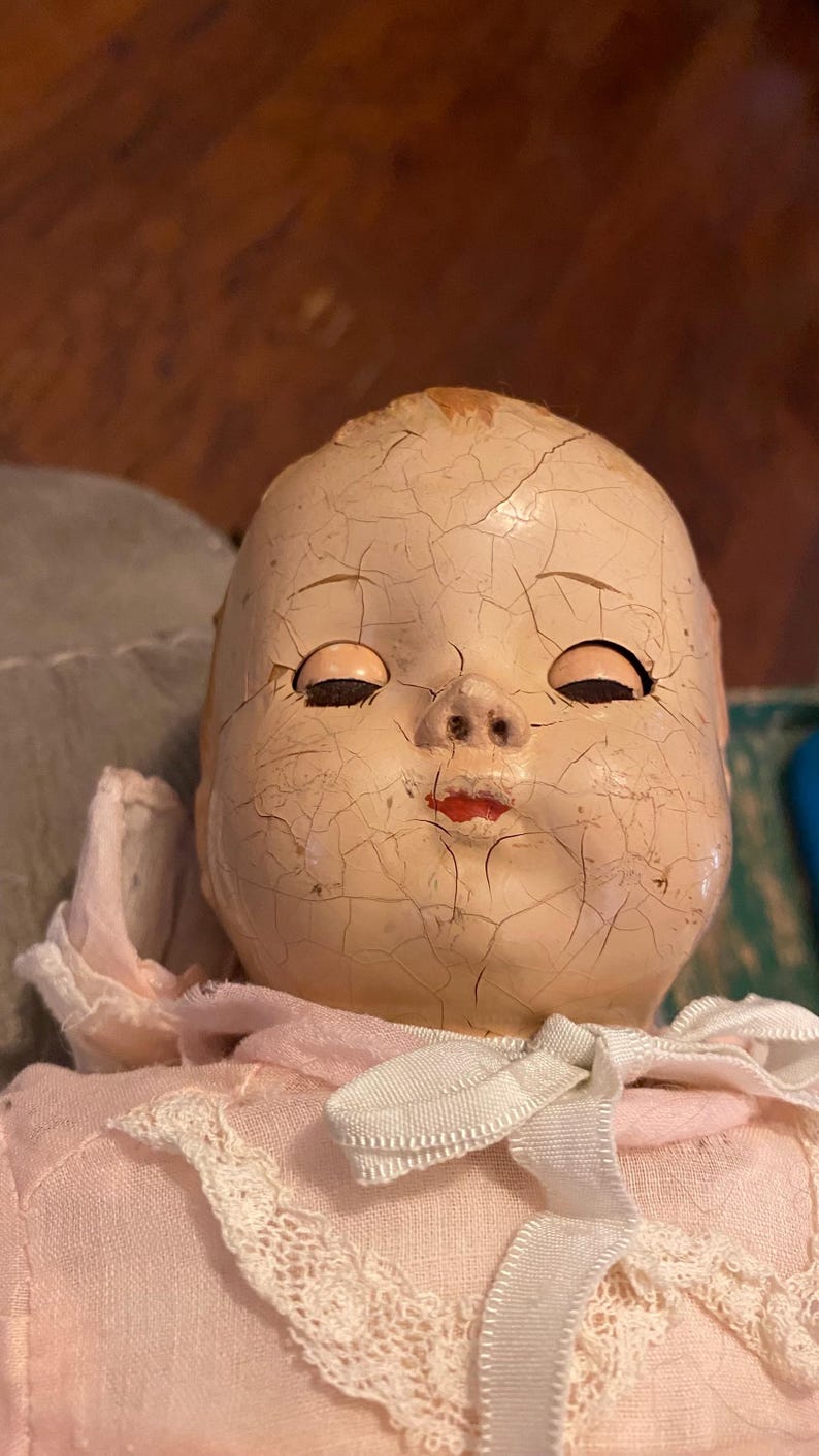 May include: A doll with a pale, cracked composition head featuring closed eyes, painted red lips, and a small nose. The doll wears a light pink fabric garment with a white lace trim collar and a tied white ribbon bow at the neck area.