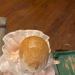 May include: A doll with a painted brown head featuring visible cracks and a textured surface. The doll wears a pale pink dress with a high, ruffled collar and white lace trim around the neckline, with a small white ribbon bow at the center.