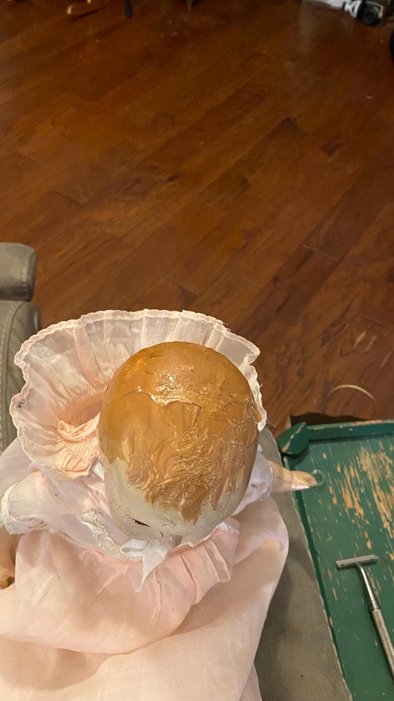 May include: A doll with a painted brown head featuring visible cracks and a textured surface. The doll wears a pale pink dress with a high, ruffled collar and white lace trim around the neckline, with a small white ribbon bow at the center.