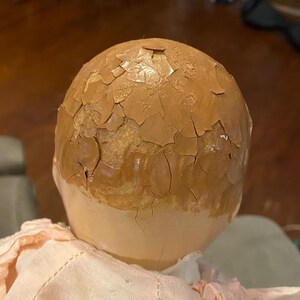 May include: A doll head with a light beige lower face and a brown upper scalp area featuring a heavily cracked and peeling paint finish. The doll is wearing a pale pink fabric garment with visible stitching around the neck and shoulder area.
