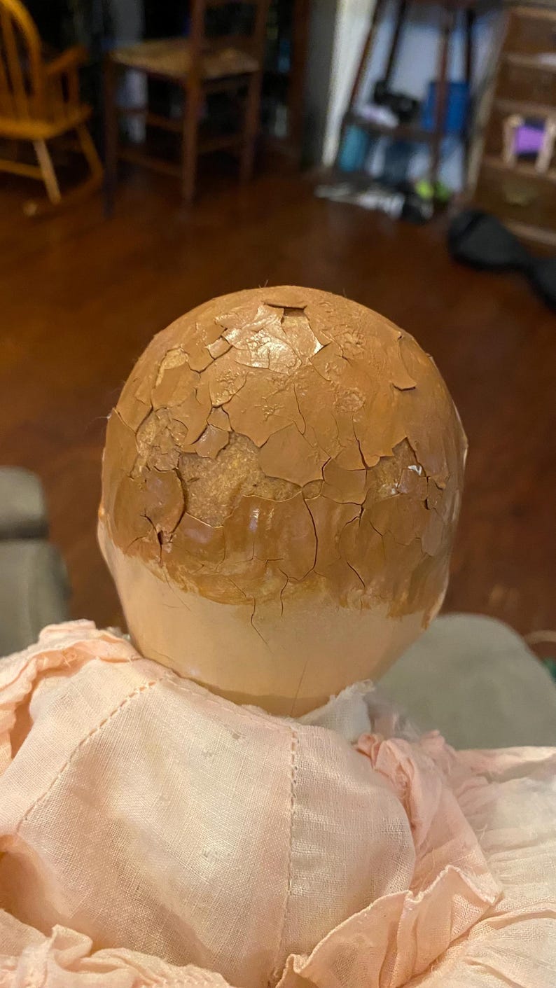 May include: A doll head with a light beige lower face and a brown upper scalp area featuring a heavily cracked and peeling paint finish. The doll is wearing a pale pink fabric garment with visible stitching around the neck and shoulder area.
