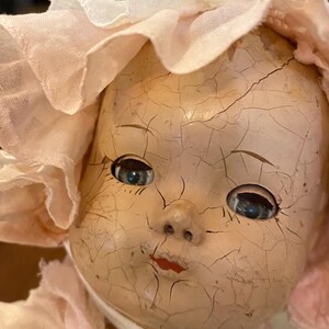 May include: A vintage doll with a heavily crazed, pale face, blue eyes, and red painted lips. The doll wears a light pink fabric bonnet with a ruffled edge and a matching pink garment featuring delicate white lace trim tied under the chin.