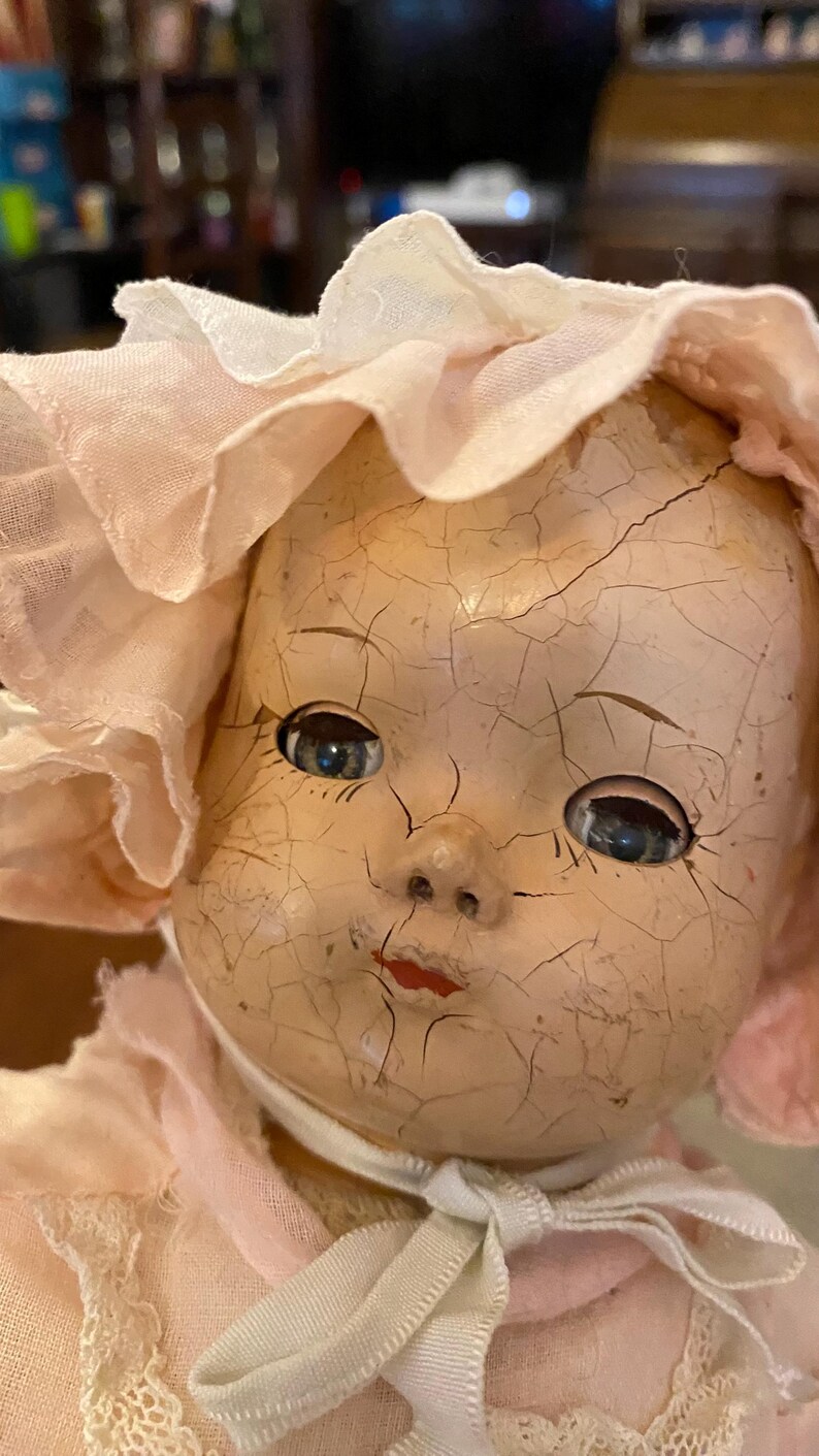 May include: A vintage doll with a heavily crazed, pale face, blue eyes, and red painted lips. The doll wears a light pink fabric bonnet with a ruffled edge and a matching pink garment featuring delicate white lace trim tied under the chin.