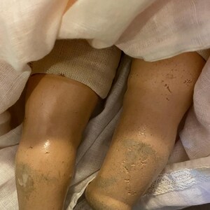 May include: A doll with light tan, textured legs showing signs of wear and discoloration, partially covered by a pale pink fabric garment featuring delicate lace trim at the hem and a soft, layered texture throughout the fabric.